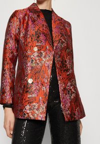 Blazer s červeným a černým vzorem s metalickými akcenty, s klopovým límcem a čtyřmi kulatými knoflíky, kombinovaný s černými flitrovými kalhotami.