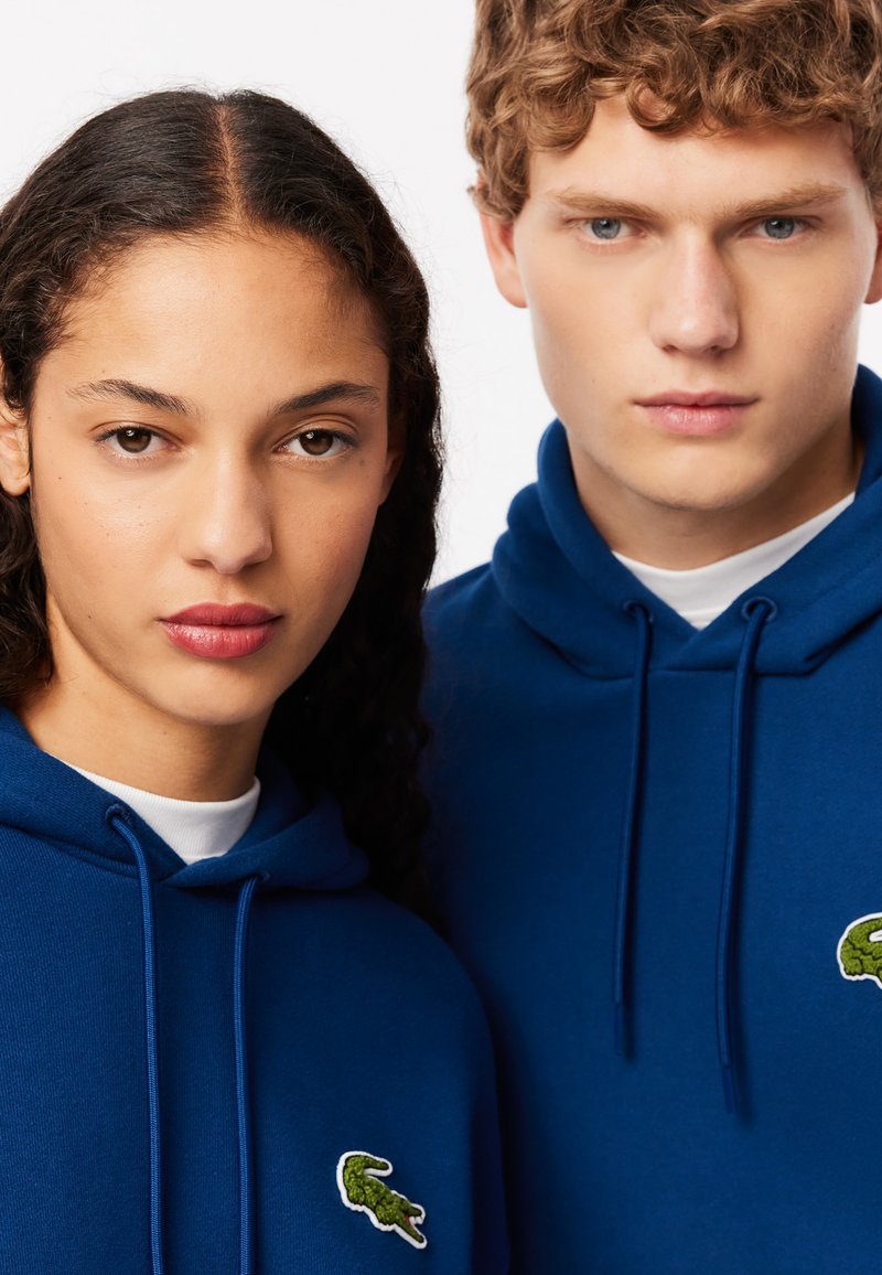 Sudaderas azul marino con cordones, que presentan logotipos de cocodrilo verde bordados en el lado izquierdo del pecho. La tela parece suave y lisa.