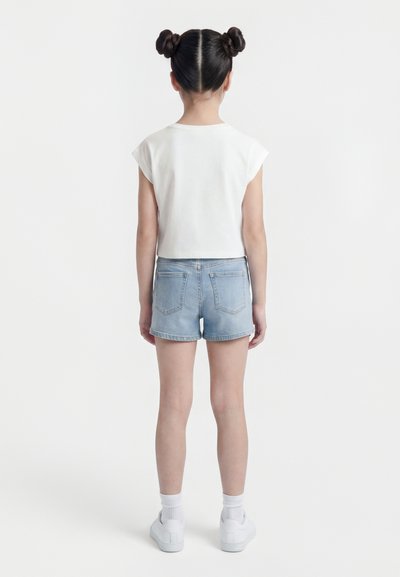 Παιδί που στέκεται πλάτη, φοράει λευκό μπλουζάκι με κοντά μανίκια, ανοιχτό μπλε τζιν σορτς, λευκές κάλτσες και λευκά αθλητικά παπούτσια, με τα μαλλιά κομμένα σε δύο κότσους.