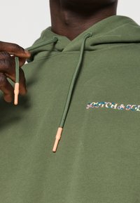 Osoba s tmavou pokožkou si upravuje zelený šňůrkový kapuci s broskvově zbarvenými konci, na prsou je vidět víceméněbarevné logo „SCOTCH & SODA“.