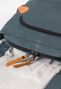 Dark blue fabric backpack with a zipper and a tan leather accent. Inside features a light grid pattern. Zipper pulls are black and tan.