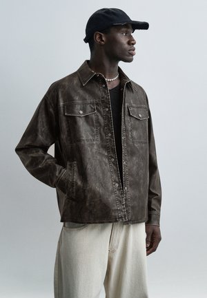 Homme portant une veste en cuir marron foncé, une casquette noire, une chemise noire, un collier de perles et un pantalon beige clair, regardant vers sa droite avec une main dans la poche.