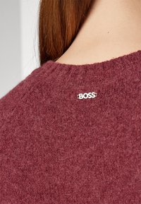 Gros plan d'une personne aux longs cheveux roux portant un pull texturé bordeaux avec un petit logo argenté « BOSS » sur le haut du dos.