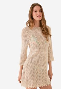 Abito a maglia beige con motivo a zigzag, dotato di maniche a tre quarti e orlo ondulato. Materiale leggero e strutturato.