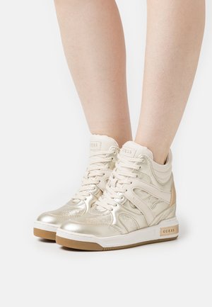Metalen gouden hoge sneakers met veters, gedragen op blote benen, voorzien van gumzolen en zichtbare branding op de tong en zijkant.