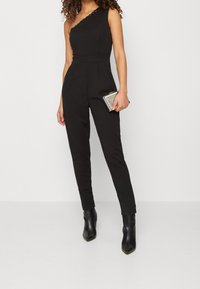 Combinaison noire à une épaule avec une silhouette ajustée, un corsage texturé, des poches latérales et des jambes fuselées. Association avec une pochette métallique.