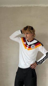 Λευκή μακρυμάνικη φανέλα ποδοσφαίρου με μαύρες, κόκκινες και κίτρινες διαγώνιες ρίγες· διαθέτει στρογγυλή λαιμόκοψη και έμβλημα στο κέντρο. Μαύρο παντελόνι.