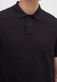Homme portant un polo noir à manches courtes avec un col, deux boutons noirs et un petit logo "BOSS" sur la poitrine gauche.
