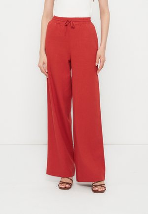 Γυναίκα με κόκκινο παντελόνι ψηλόμεσο, φαρδύ με κορδόνι στη μέση και καφέ σανδάλια με λουριά, στέκεται σε λευκό φόντο.