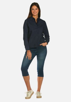 Chemise boutonnée marine, design ajusté, associée à des capris en denim foncé de longueur mi-mollet. Baskets beiges à texture légère. Le mannequin se tient de manière décontractée.