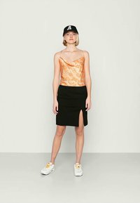 Top de alças com padrão laranja e decote drapeado, combinado com uma saia preta com uma fenda lateral. O modelo usa ténis brancos e uma camisola preta.