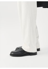 Mocassins en cuir noir portés avec un pantalon blanc sur mesure, montrés de profil sur un fond clair uni.