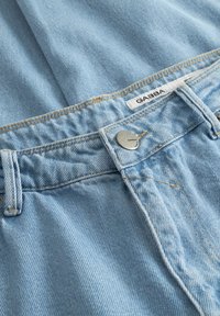 Lys blå denimjeans nærbilde som viser midjebånd, beltehemper, metallknapp med "Gabba" merke, og en hvit tekstillapp.