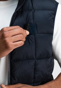 Marinefarbener Puffer-Weste mit Steppdetails und Reißverschluss. DieTrägerin passt ein strukturiertes, quadratisches Klettpatch im Brustbereich an.