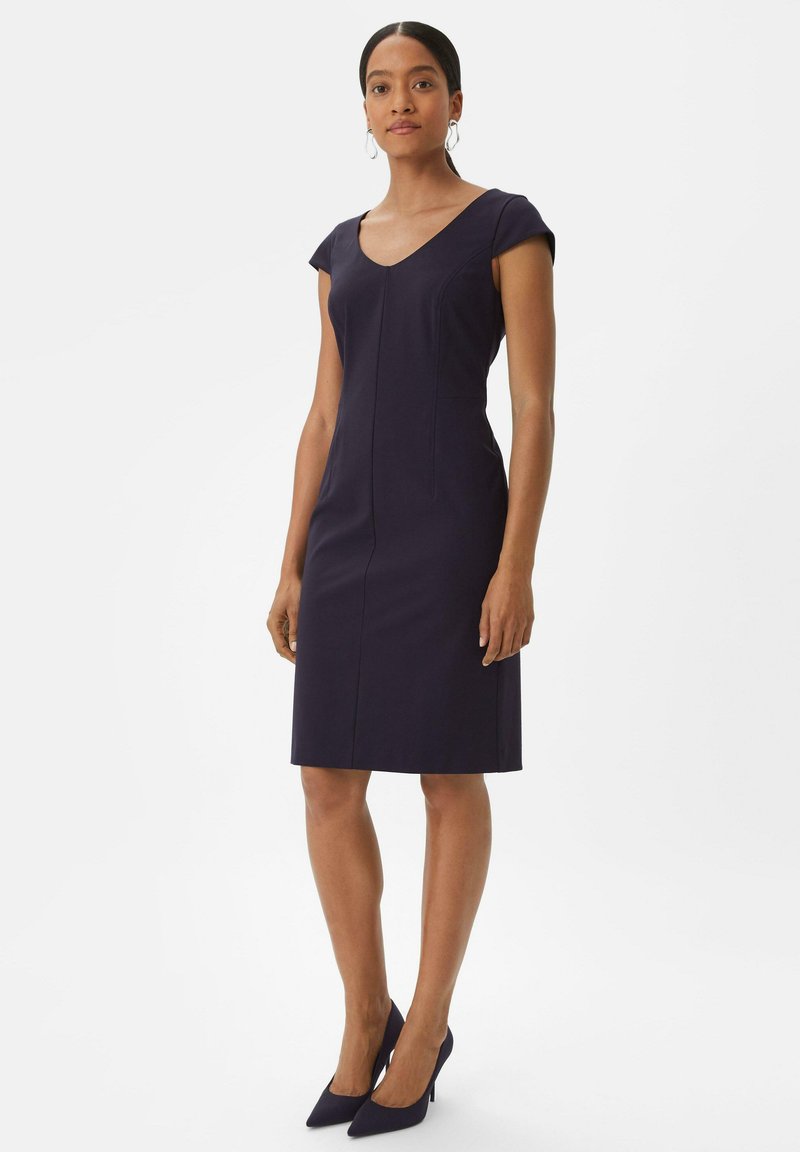 Marineblaues, tailliertes Kleid mit kurzen Cap-Ärmeln, V-Ausschnitt und vertikalen Nähten, kombiniert mit passenden spitzen Absätzen. Glatte Stoffoberfläche.