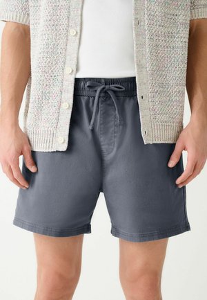 Mann trägt marineblaue Shorts mit Kordelzug, weißes ärmelloses Shirt und hellgraue Strickjacke mit Knöpfen, Hände entspannt an den Seiten.