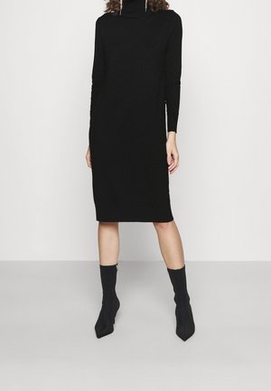 Femme portant une robe noire à manches longues, longueur genoux, col roulé et des bottines noires à talons, debout devant un fond uni.