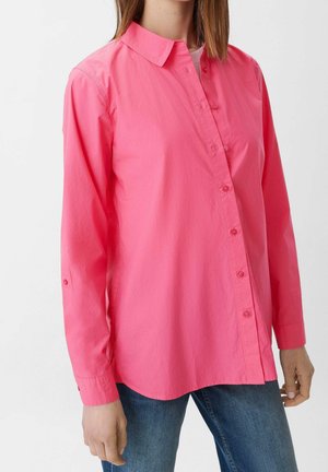 Femme portant une chemise rose vif à manches longues avec col, associée à un jean en denim bleu, sur un fond uni.