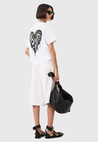 Hvid bomulds T-shirt med sort drippende hjerte grafik på ryggen, parret med hvide shorts og sorte sandaler. Accessoriseret med en sort taske.