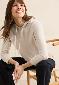 Felpa con cappuccio grigio chiaro dall'aspetto texture, con tasca a marsupio frontale e chiusura con cordino, abbinata a jeans in denim scuro.