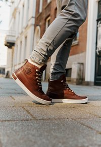 Zapatillas altas de cuero marrón con suelas blancas, que cuentan con ojales plateados, un logo circular en el lateral y detalles de cuero texturizado.