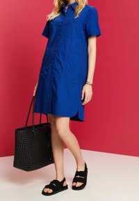 Donna che indossa un vestito blu con bottoni e sandali neri, mentre tiene una grande borsa a tracolla nera intrecciata su uno sfondo rosso e bianco.