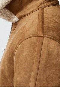 Brun ruskindsjakke med en tekstureret overflade, der har en hætte foret med lammeskind og lynlås, med fremhævede bløde syningsdetaljer.