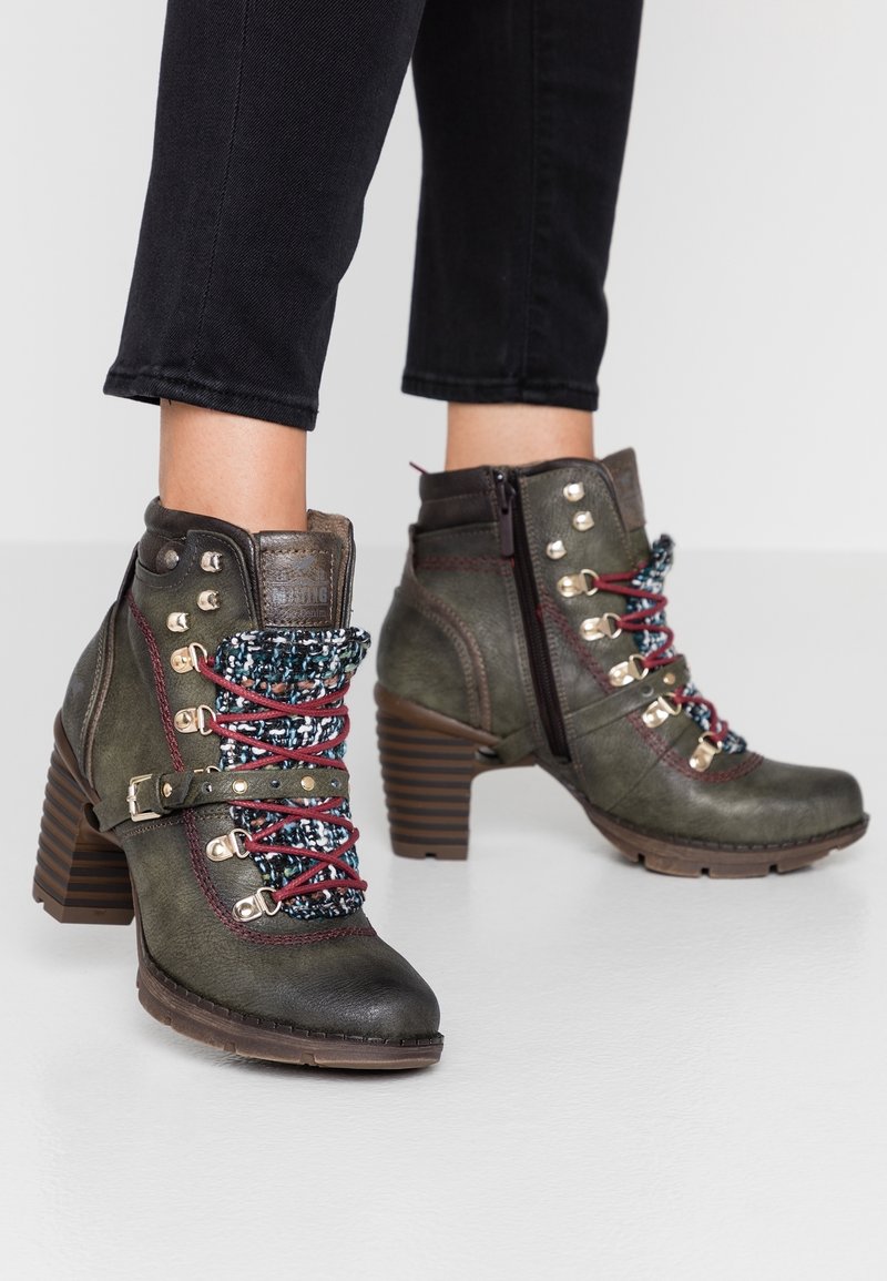 Botas de cuero verde oscuro hasta el tobillo con cordones burdeos, lengüeta de tela con patrón, cremallera lateral, correa con hebilla y tacones gruesos marrones, usadas con pantalones negros.