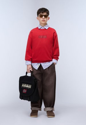 Chlapec s slunečními brýlemi, červená mikina přes proužkovanou košili, hnědé ohrnuté kalhoty, hnědé boty, drží černý batoh s norskou vlajkou na nášivce.