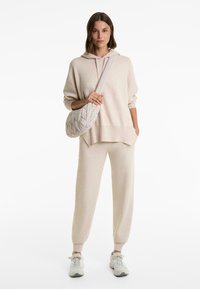 Woman wearing beige knit hoodie and pants with white sneakers, carrying a padded beige crossbody bag, standing on white background.