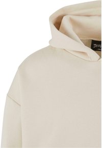 Lys beige hettegenser med en avslappet passform, med snøring på hetta og myk tekstur. Minimalistisk design uten synlige mønstre.