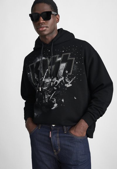Felpa nera con stampa grafica che presenta il logo "KISS" e i membri della band, abbinata a jeans scuri. Accompagnata da occhiali da sole angolari.