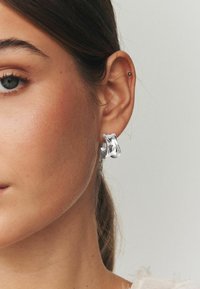 Sudraba stīpas auskaru ar viļņotu dizainu, gludu tekstūru un pulētu virsmu, valkā uz auss gaišā fonā.