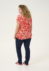 Rød blomsterbluse med hvide blomster, korte ærmer og en afslappet pasform, parret med mørkeblå jeans og røde sandaler. Glat tekstur.