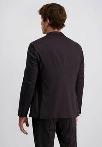 Mann mit dem Rücken zur Kamera, der einen dunklen Sakkos und eine dunkle Hose trägt, vor einem einfarbigen hellen Hintergrund.