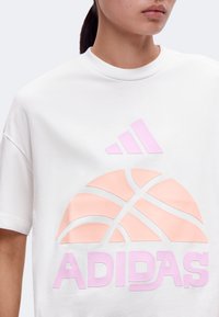 Άτομο που φοράει λευκό μπλουζάκι Adidas με ροζ γραφικό καλαθοσφαίρισης και το λογότυπο Adidas σε ροζ χρώμα στο μπροστινό μέρος.