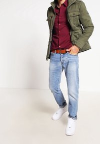 Persoon in een groene jas, geruite rode shirt, lichtblauwe jeans en witte sneakers, staand tegen een effen witte achtergrond.