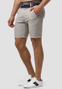 Graue Baumwollshorts mit umgeschlagenen Säumen, Seitentaschen und einem gewebten marineblauen Gürtel mit braunem Lederakzent. Weiße Sneaker mit schwarzen Details.