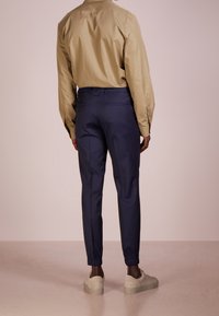 Beige långärmad skjorta i kombination med navy tapered byxor med elastiska muddar och grå sneakers, sedd bakifrån.