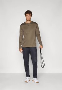 Fiatal férfi olíva zöld, hosszú ujjú pólóban, fekete sportnadrágban és fehér tornacipőben, fekete ugrókötéllel áll egy sima, világos háttér előtt.