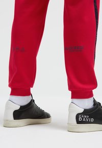 Rote Jogginghose mit marineblauen seitlichen Streifen, mit grafischen Drucken und elastischen Bündchen. Schwarz-weiße Sneakers mit "CAMP DAVID" Branding.