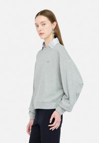 Sweatshirt gris avec une coupe décontractée, des manches à revers et un logo brodé. Associé à une chemise à col avec un col à motif.
