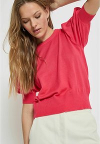Pull rouge à manches courtes avec un ourlet et des poignets côtelés, présentant une coupe décontractée. Texture lisse, design simple et sans motifs visibles.