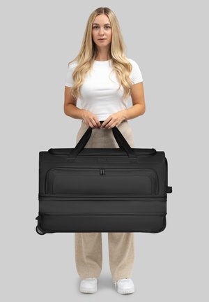 Donna con lunghi capelli biondi che indossa una maglietta bianca e pantaloni beige, mentre tiene una grande valigia nera con ruote per le maniglie.