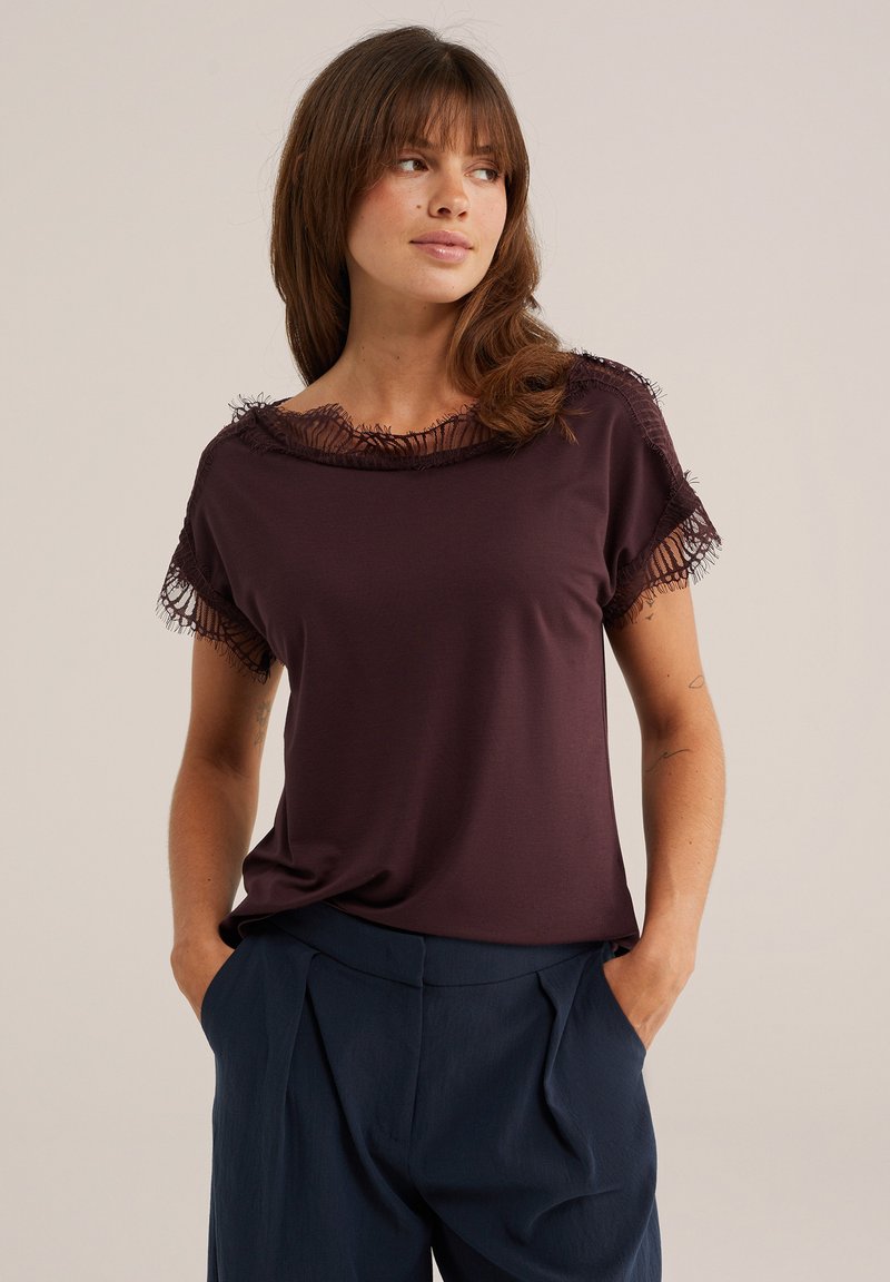 Femme aux cheveux bruns portant un haut à manches courtes violet foncé avec bordure en dentelle et un pantalon bleu marine, debout les mains dans les poches, regardant sur le côté.