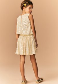 Roomkleurige bloemenpailletten jurk met een mouwloze top, elastische tailleband en gelaagde rok; gecombineerd met gouden ballerina's.