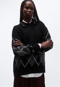 Maglione nero lavorato a maglia con scollo con colletto, presenta un motivo a rombi grigio e bianco sulle maniche e polsini a costine.