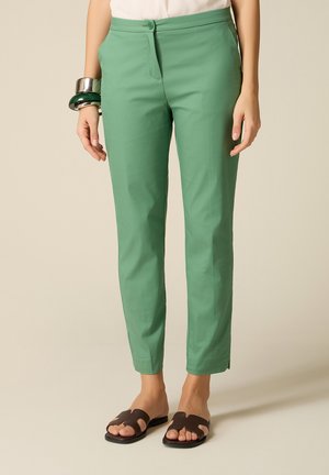 Donna che indossa pantaloni verdi aderenti alla caviglia, top bianco, sandali neri con punta aperta e bracciali spessi argentati e verdi al polso sinistro.