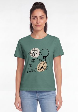 Frau mit dunklen Haaren in einem grünen T-Shirt mit einer schwarzen Linienzeichnung eines Gesichts mit Blumen und dem Wort "love,", kombiniert mit hellblauen Jeans.
