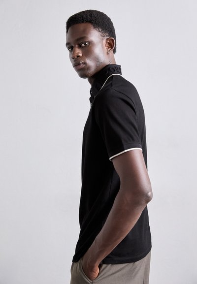 Tricou polo negru cu detalii albe, având un guler texturat marcat cu "EMPOWER", purtat cu pantaloni gri. Modelul este văzut din profil.